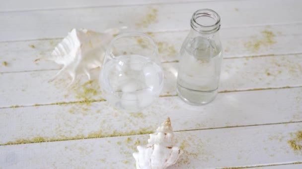 Verre avec eau et coquillages blancs sur table en bois avec sable — Video