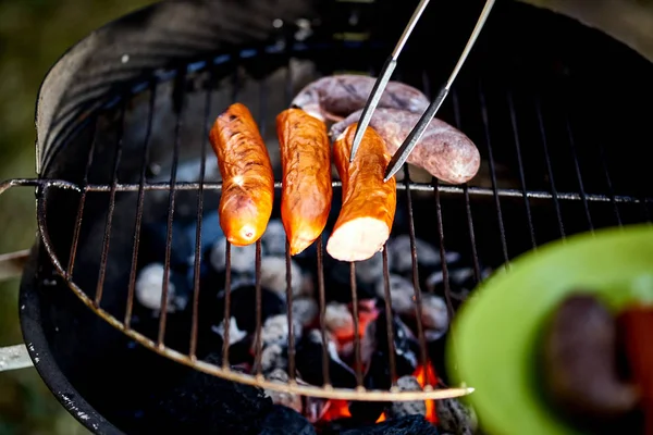 Soczysta kiełbasa na gorącym grillu z szczypce podczas letniego grilla — Zdjęcie stockowe