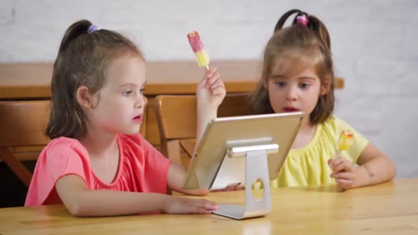 Две девчонки едят мороженое и смотрят мультфильм на столе — стоковое видео