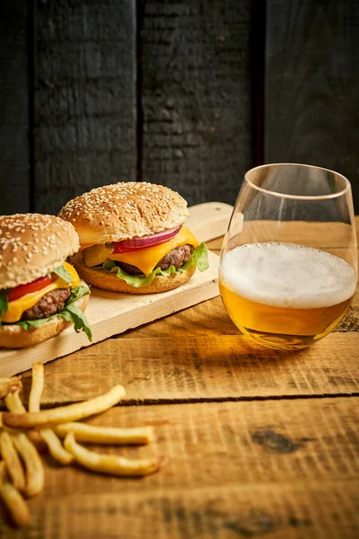 Two tasty burgers and fries on wooden board and black background — Stock Photo, Image