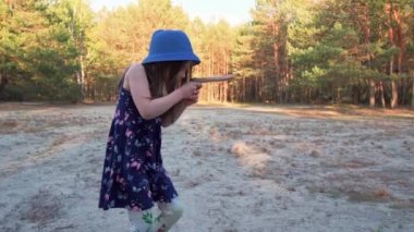 Yazlık elbiseli küçük kız çam ormanında tahta bir oyuncak silah tutuyor.