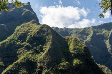 Parçalı bulutlu bir gökyüzünün altında yoğun bitki örtüsüyle kaplı sarp yeşil dağ yamaçları engebeli volkanik araziyi ve Endonezya 'nın doğal güzelliğini gözler önüne seriyor..