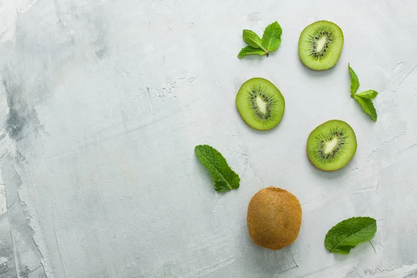 Fresh kiwi whole fruit and three kiwi slices with mint leaves on grey concrete background diagonally. Creative layout, food concept, flat lay, top view, horizontal orientation, place for copy space.