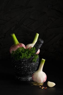 Mermer harç ve dibekler ile yeşil otlar genç sarımsak ampuller, diş sarımsak, koyu arka plan üzerine biber. Karanlık anahtar aydınlatma. Dikey yönlendirme, yer boşaltmak için.