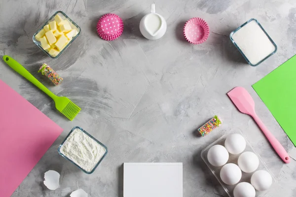 Cooking baking for kids flat lay background concept. Pink green paper white notebook kitchen tools silicone brush spatula colored sprinkling eggs sugar flour butter cupcake molds milk jug concrete.