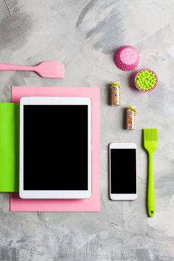 Tablet cep telefonu uygulama hizmeti Web sitesi mockup mutfak tereyağı un şeker yumurta pişirme silikon fırça pembe spatula kek kalıpları yeşil yağmurlama gri beton araçları. Üstten görünüm düz yatıyordu.