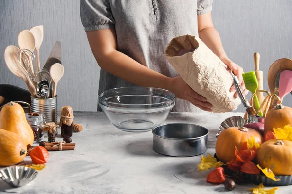 Woman cooking baking pack of flour pumpkins autumn time. Glass plate, sieve, pack of flour, physalis, kitchen tools, spices, leaves, gray background.