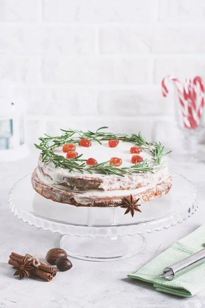 Christmas cake on glass stand decorated with rosemary and dried red berries. Winter holiday composition with candy canes, bottle of milk, candle lamp and fir-trees, colored sprinkling.