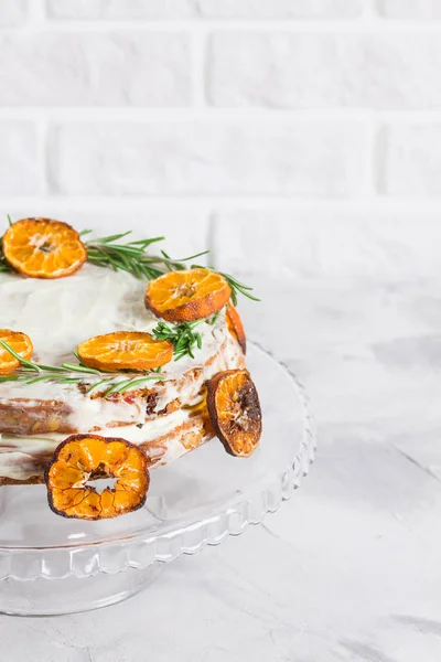 Cake on glass stand decorated with rosemary and dried mandarin slices on grey concrete white brick wall background. Close up, vertical orientation, place for copy space.