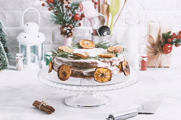 Christmas cake on glass stand decorated with rosemary and dried mandarin slices. Winter holiday composition cinnamon, gift box, metallic spoon, kitchen stuff tools.