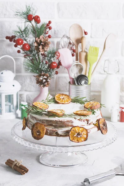 Christmas cake on glass stand decorated with rosemary and dried mandarin slices. Winter holiday composition cinnamon, gift box, metallic spoon, kitchen stuff tools.