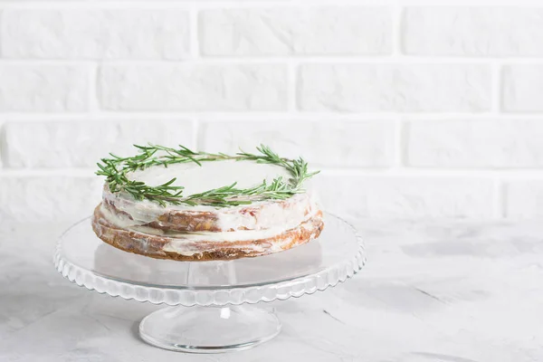 Cake on glass stand decorated with sprigs of rosemary on grey concrete white brick wall background. Horizontal orientation, place for copy space.