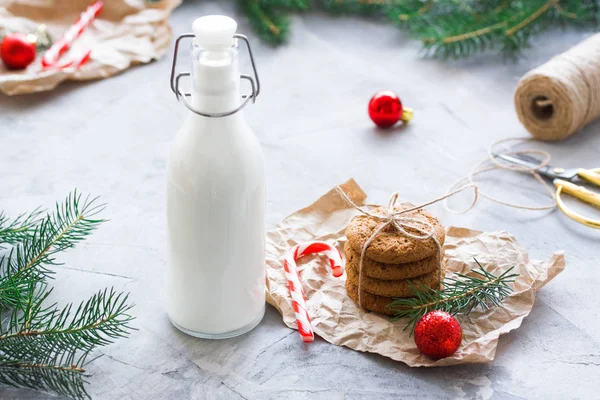 Milk in glass bottle and oatmeal cookies tied with string in craft paper for Santa Claus, skein of thread, candy canes, balls, fir-trees branches, gray concrete background. Winter holiday concept.