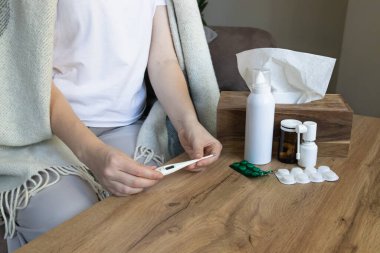 Hasta kadın battaniyeye sarılı olarak kanepeye oturmuş vücut ısısını ölçmek için termometre kullanıyor. Soğuk algınlığı, ateş ve mevsimsel hastalık kavramı.