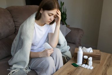 Kanepede başı ağrıyan hasta bir kadın, ateşi, viral enfeksiyonu ve mevsimsel soğuk yüzünden bitkin ve bitkin hissediyor. Soğuk algınlığı, grip, kişisel bakım ve evde tedavi kavramı.