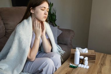 Elleri boğazında, boğaz ağrısı ve ateşi olan hasta bir kadın battaniyeye sarılmış oturuyor. Grip, soğuk algınlığı, anjin, farenjit ve sağlık tedavisi.