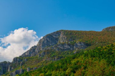 Kayalık uçurumlar ve sonbahar renklerini gösteren orman yamaçlarıyla manzaralı bir dağ manzarası, açık mavi gökyüzü altında yumuşak beyaz bulutlar, manzara ve seyahat temaları için ideal..
