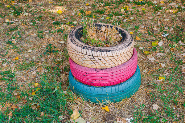 Three old car tires painted in beige, pink and blue are stacked outdoors and repurposed as a garden planter. Grass and dry leaves fill the top tire on a patchy lawn background.