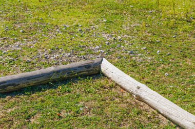 Bir açıya yerleştirilmiş iki aşınmış ahşap kütük küçük kayalarla yeşil çimlerde kırsal bir sınır oluşturarak basitlik, yapı ve doğal doğa elementlerini çağrıştırıyor..