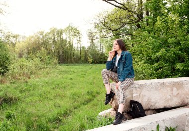Uzun kahverengi saçlı genç kız yeşil yaz, bahar ormanlarında taşın üzerinde oturur. Metin için yer.