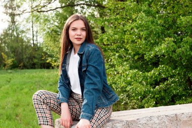 Uzun kahverengi saçlı genç kız yeşil yaz ve bahar ormanlarında taşın üzerinde oturuyor..