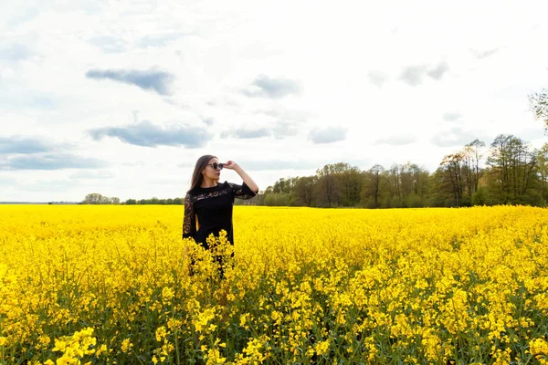 Krásná hnědovlasá dívka v slunečních brýlích s dlouhými vlasy v černých šatech stojí ve žlutém poli. — Stock fotografie