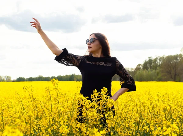 Krásná hnědovlasá dívka ve slunečních brýlích s dlouhými vlasy v černých šatech stojí na žlutém poli. Koncept léta, mládí, svoboda. — Stock fotografie