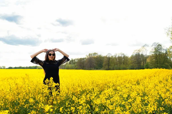 Krásná hnědovlasá dívka ve slunečních brýlích s dlouhými vlasy v černých šatech stojí na žlutém poli. Koncept léta, mládí, svoboda. — Stock fotografie