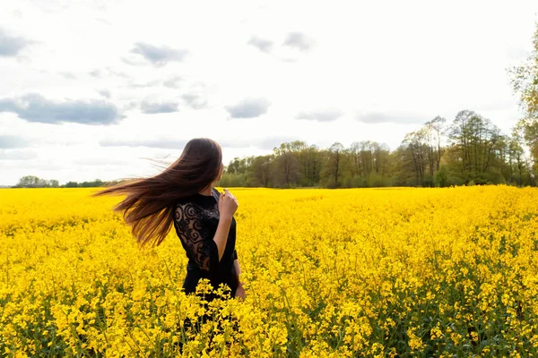Krásná hnědovlasá dívka s dlouhými vlasy v černých šatech stojí na žlutém poli. Koncept léta, mládí, svoboda. — Stock fotografie
