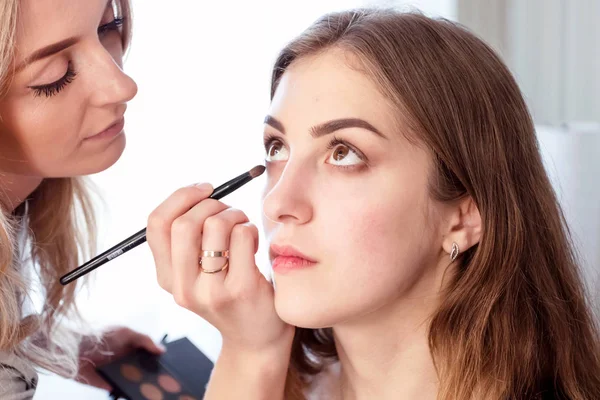 O artista de maquilagem faz a maquilagem da tarde da menina jovem de cabelos castanhos. Roupa casual. Backstage do processo. — Fotografia de Stock