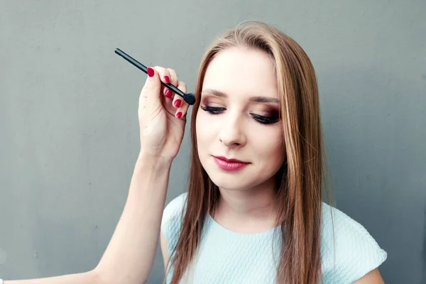 Schöne junge Mädchen mit professionellem Make-up auf grauem Hintergrund. Maskenbildnerin macht Augen-Make-up. — Stockfoto