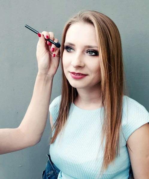 Schöne junge Mädchen mit professionellem Make-up auf grauem Hintergrund. Maskenbildnerin macht Augen-Make-up. — Stockfoto