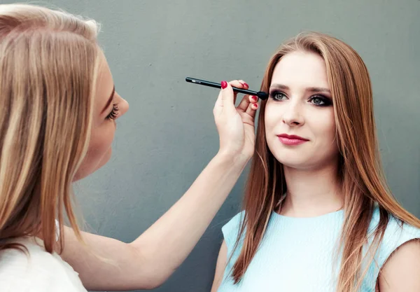 Schöne junge Mädchen mit professionellem Make-up auf grauem Hintergrund. Maskenbildnerin macht Augen-Make-up. — Stockfoto