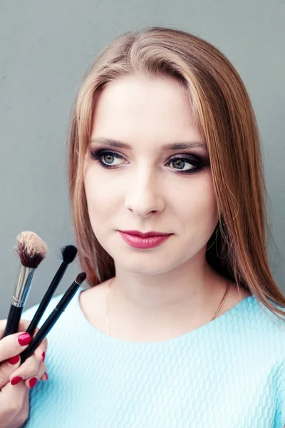 Krásná mladá dívka s profesionální make-up a make-up štětce na šedém pozadí. Koncept vzhazhist, móda, kosmetologie. — Stock fotografie