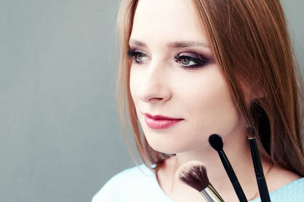 Schöne junge Mädchen mit professionellem Make-up und Make-up-Pinsel auf grauem Hintergrund. Konzept vzhazhist, Mode, Kosmetologie. — Stockfoto
