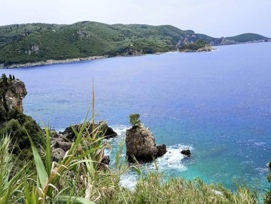 Paleokastritsa, Korfu 'daki kayalık kıyı şeridi, turkuaz İyon Denizi ve uzak yeşil tepeleri gösteren uçurumlardan görüntü. Akdeniz geçmişi veya kıyı tasarımı için temiz doğal ortam ideali.
