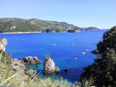 Yunanistan 'ın Korfu kentinde yemyeşil tepeleri ve kayalık kıyı şeridi olan açık mavi İyon Denizi' ndeki küçük tekneler. Kıyı bitkileri tarafından çerçevelenmiş görüntü. Seyahat, Akdeniz veya yaz tasarım kullanımı için ideal.
