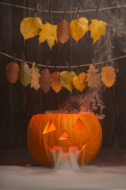 Cadılar Bayramı balkabağı oyulmuş, parlak yüzlü ve dumanlı, yukarıda sarkan canlı sonbahar yapraklarıyla çevrili, kutlamalar için neşeli ve ürkütücü bir ortam yaratıyor.