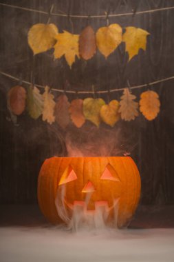 Parlayan yüzlü Cadılar Bayramı balkabağı, canlı sonbahar yapraklarının üzerine kurulmuş dumanlar salıyor, mevsimsel şenlikler için festival ortamını güçlendiriyor.