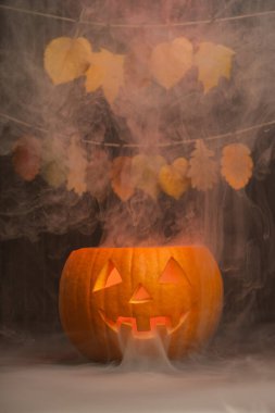 Kıvrımlı duman ve sonbahar yapraklarıyla çevrili parlayan balkabağı feneri Cadılar Bayramı ve mevsimsel dekorasyonların ürkütücü atmosferini güçlendiriyor.