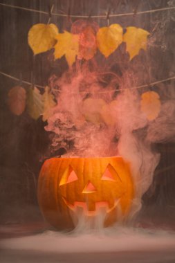 Oyulmuş Cadılar Bayramı balkabağı dumanı yayar, yukarıda sarkan canlı sonbahar yapraklarıyla çevrili, mevsimsel kutlamalar için neşeli ve ürkütücü bir ortam yaratır.