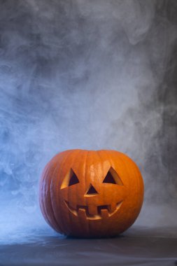 Yüzü gülümseyen turuncu balkabağı dumanla kaplanmış karanlık bir yüzeyde duruyor. Bu da bayram ruhunu ve mevsimsel kutlamaları çağrıştırıyor.