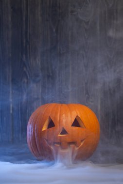 Gülümseyen bir yüzle oyulmuş Cadılar Bayramı balkabağı, sis ve dumanla kaplanmış, karanlık ahşap bir zemin üzerinde, festival sezonunun ürkütücü atmosferini güçlendiriyor.