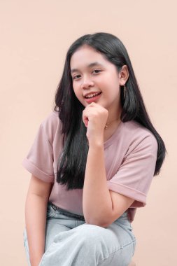 Modern tarzda giyinmiş Asyalı bir kadın, pastel bir tişört ve kot pantolon giyiyor, elleri çenesinde gülümsüyor, kendine güvenini, çekiciliğini, doğal güzelliğini ve gençlik ifadesini gösteriyor..