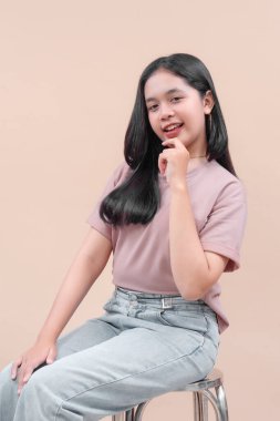 Uzun siyah saçlı, gündelik modern kıyafetli güzel bir Asyalı kadın pastel tişört ve kot pantolon giyiyor. Elleri dudaklarının yanında oturuyor ve gülümsüyor. Cazibesini, özgüvenini ve gençlik enerjisini gösteriyor..