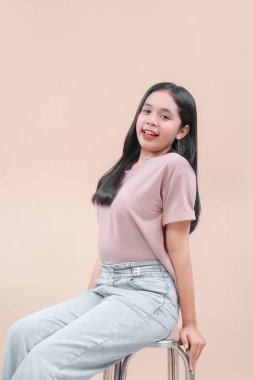 Sıradan modern tarzda Asyalı bir kadın, taburede oturuyor, pastel bir gömlek ve açık renk kot giyiyor, rahat bir şekilde gülümsüyor..