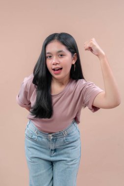 Gündelik modern kıyafetli Asyalı kadın, kendine güvenen bir gülümsemeyle poz veriyor, esnek bir jest yaparak kolunu kaldırıyor, pastel tişört ve kot pantolon giyiyor, gücü, enerjiyi ve pozitifliği simgeliyor..