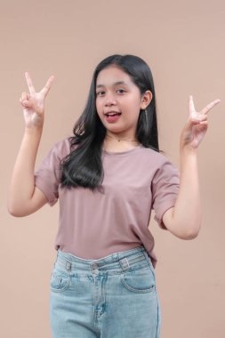Uzun siyah saçlı, günlük pembe tişört ve kot pantolon giyen Asyalı bir kadın, güvenli bir şekilde gülümserken iki elini de neşeli bir barış işareti jesti ile kaldırıyor..