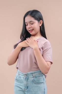 Uzun siyah saçlı, günlük tişört ve kot pantolon giyen Asyalı genç bir kadın. İki elini göğsüne koyup hafifçe gülümsüyor. Minnettarlığını, nezaketini ve samimiyetini bej arka planda gösteriyor..