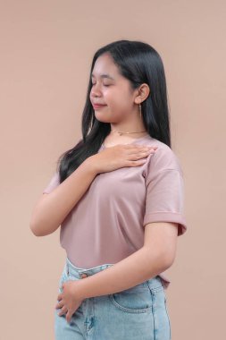 Uzun siyah saçlı, günlük tişört ve kot pantolon giyen, eli göğsünde, elleri kapalı, barışçıl şükran ve bej arka planda sakin bir güven ifade eden Asyalı genç bir kadın..